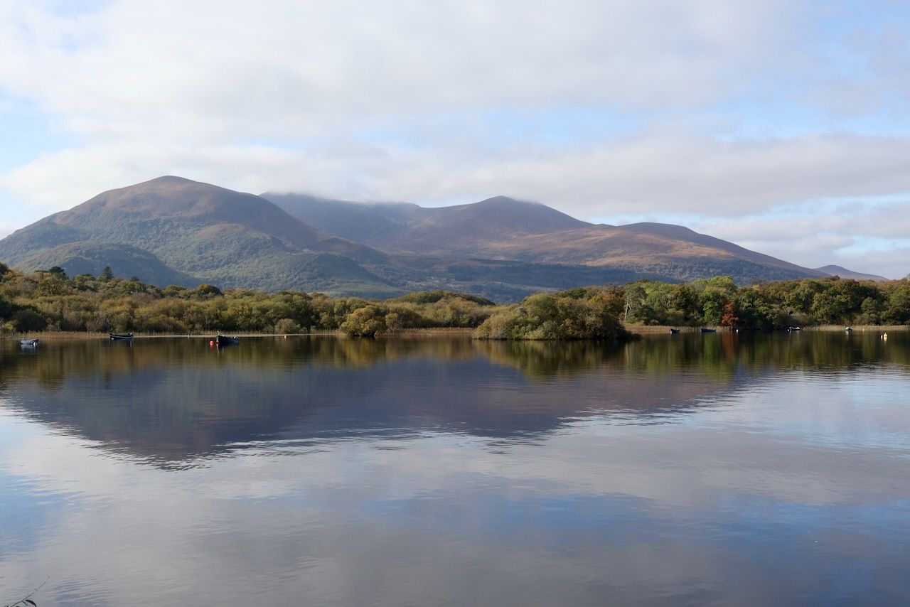 A calm lake reflecting a range of softly rounded mountains under a bright, partly cloudy sky. The shoreline is lined with dense green and autumn-tinged trees, and a few small boats sit still on the water. The mountains in the background rise gently with patches of shadow and sunlight, creating a peaceful, expansive landscape.
